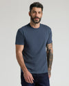 A man wearing a dark heather blue crewneck t-shirt and dark jeans, standing against a light gray studio background.