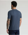 A man viewed from behind wearing a heathered slate blue short-sleeve t-shirt and dark navy jeans, standing against a light gray studio background.