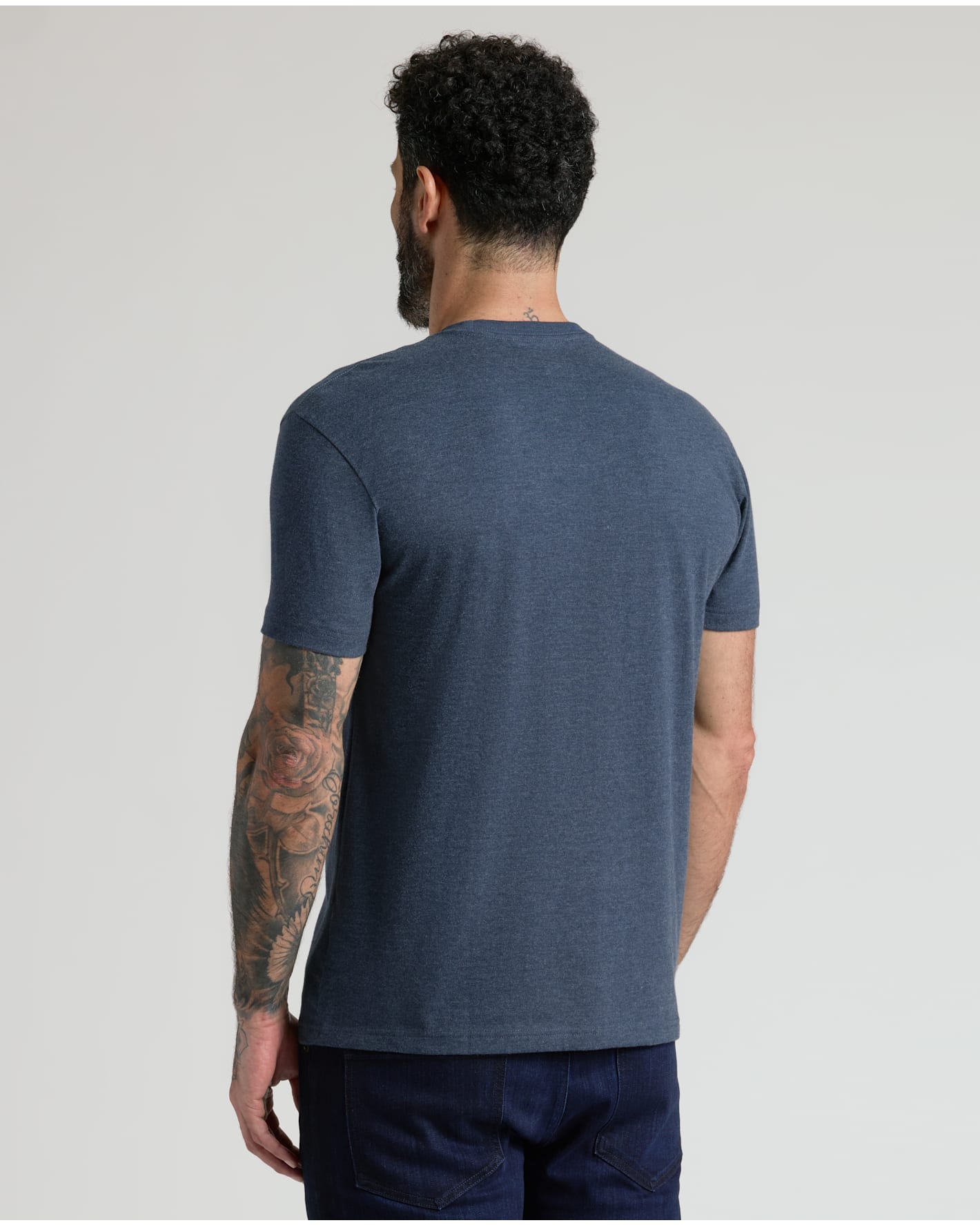 A man viewed from behind wearing a heathered slate blue short-sleeve t-shirt and dark navy jeans, standing against a light gray studio background.