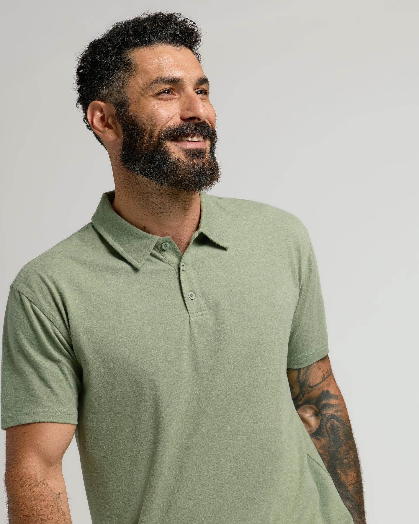 Man in Dark Heather Bay Green Classic Polo, smiling with tattoo visible.