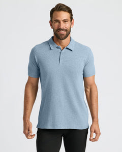 True ClassicA man wearing a light blue heathered short-sleeve polo shirt, paired with black pants, standing against a solid light gray studio background.