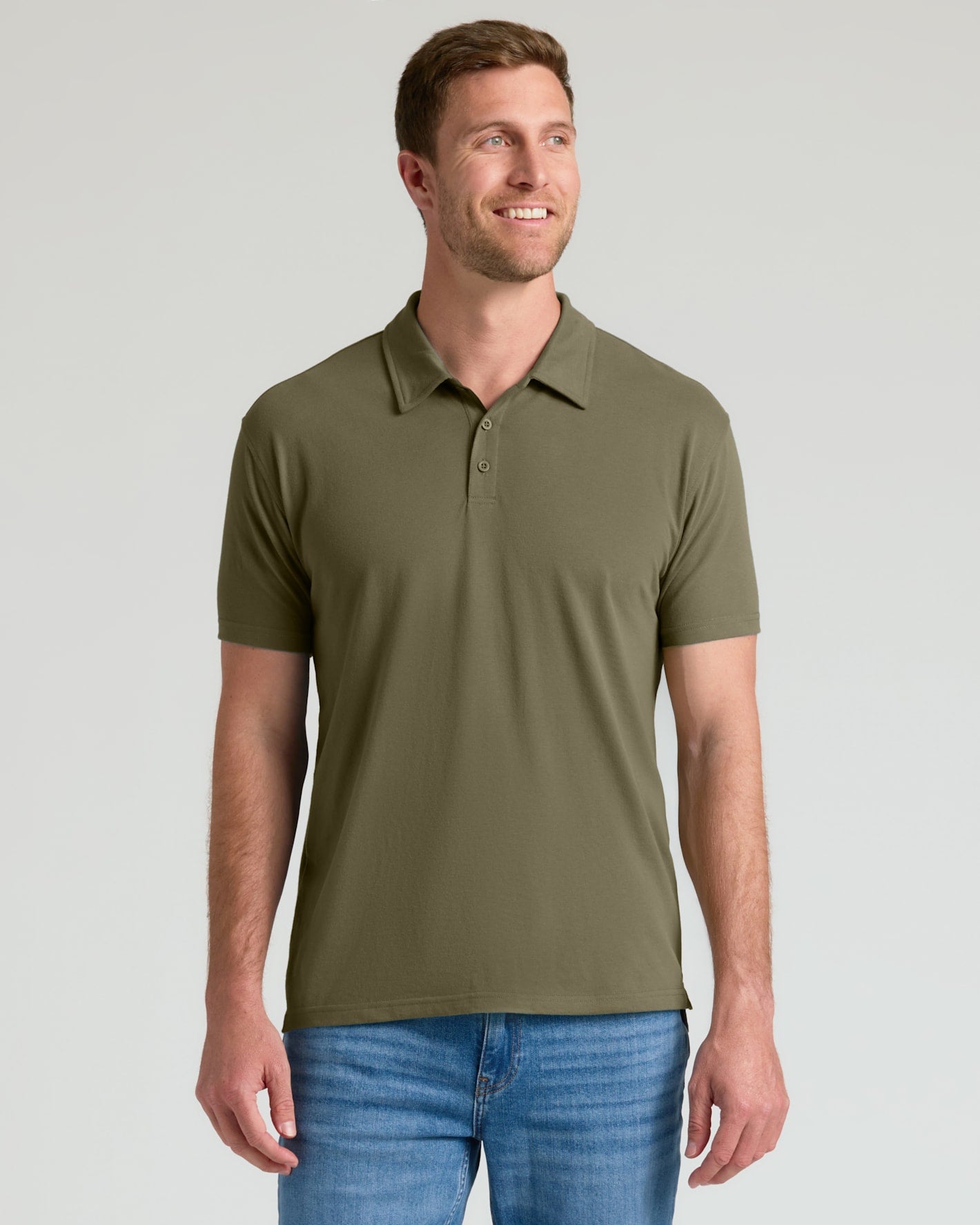A man wearing an olive green short-sleeve polo shirt and blue jeans, standing against a light gray studio background.