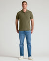 A man wearing an olive green short-sleeve polo shirt, blue jeans, and white sneakers, standing against a light gray studio background.