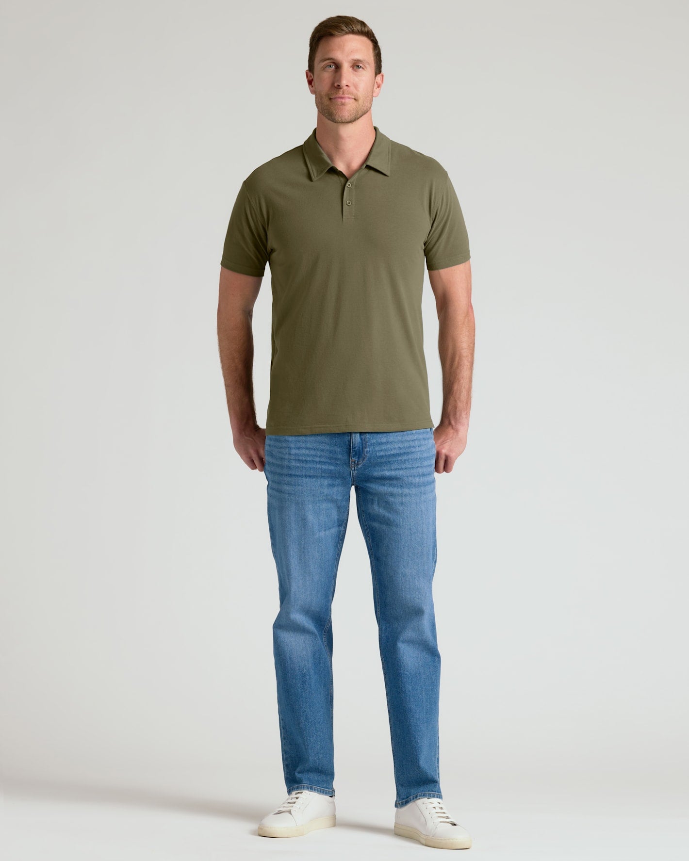 A man wearing an olive green short-sleeve polo shirt, blue jeans, and white sneakers, standing against a light gray studio background.