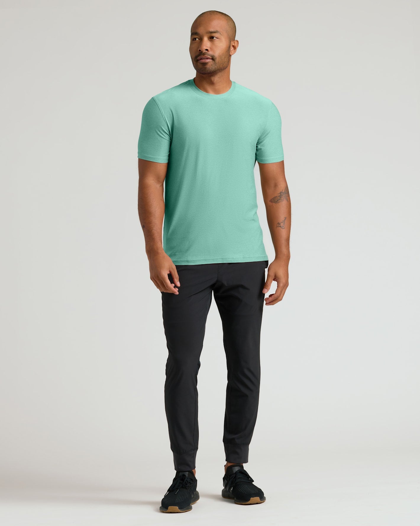 A man modeling a short-sleeve crewneck t-shirt in mint green, paired with black joggers and black sneakers, standing against a light gray studio background.
