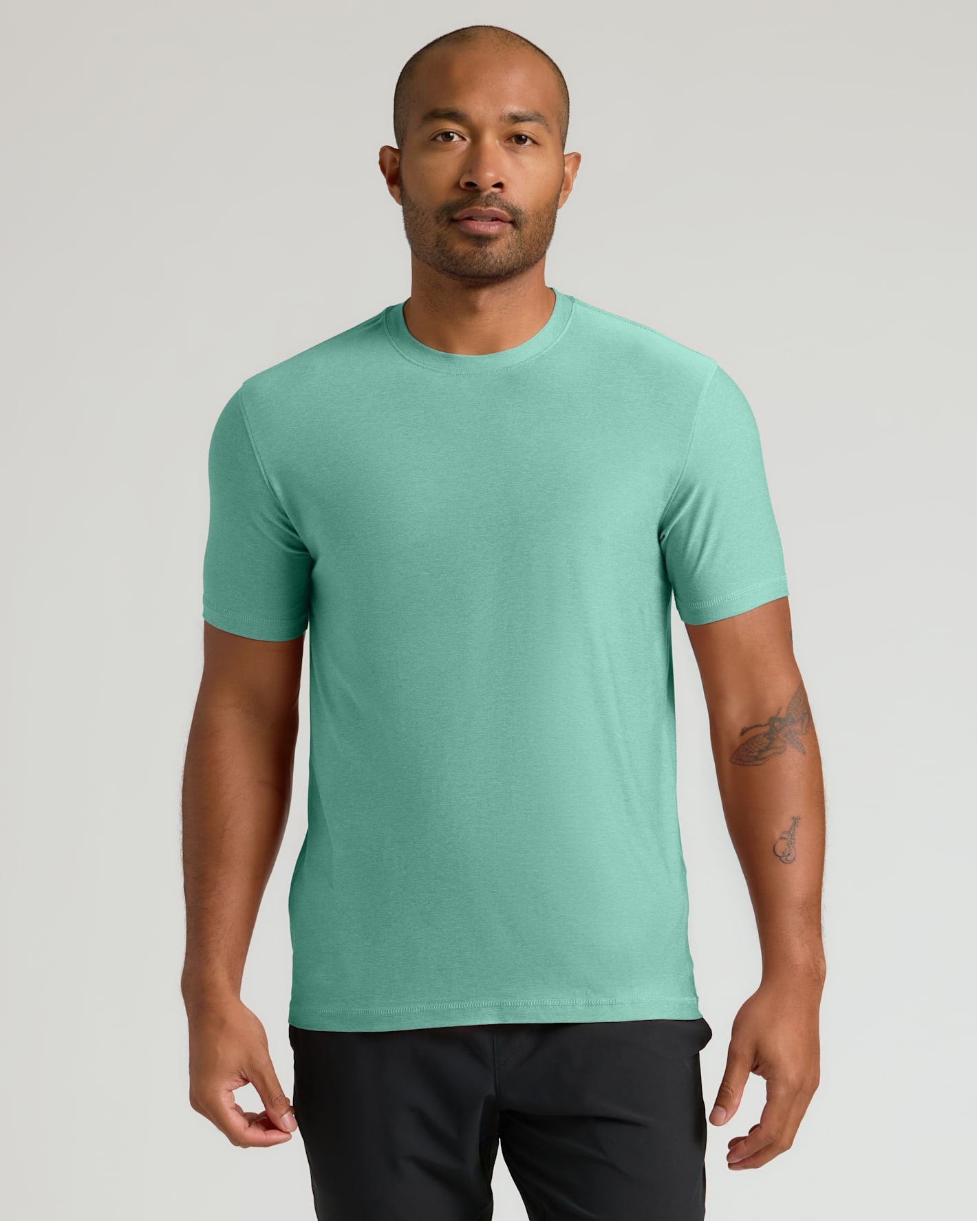 A man wearing a short-sleeve crewneck t-shirt in a solid mint green color, paired with black pants, standing against a light gray studio background.