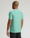 A man viewed from the back wearing a short-sleeve crewneck t-shirt in a heathered mint green color, paired with black athletic pants, against a light gray studio background.