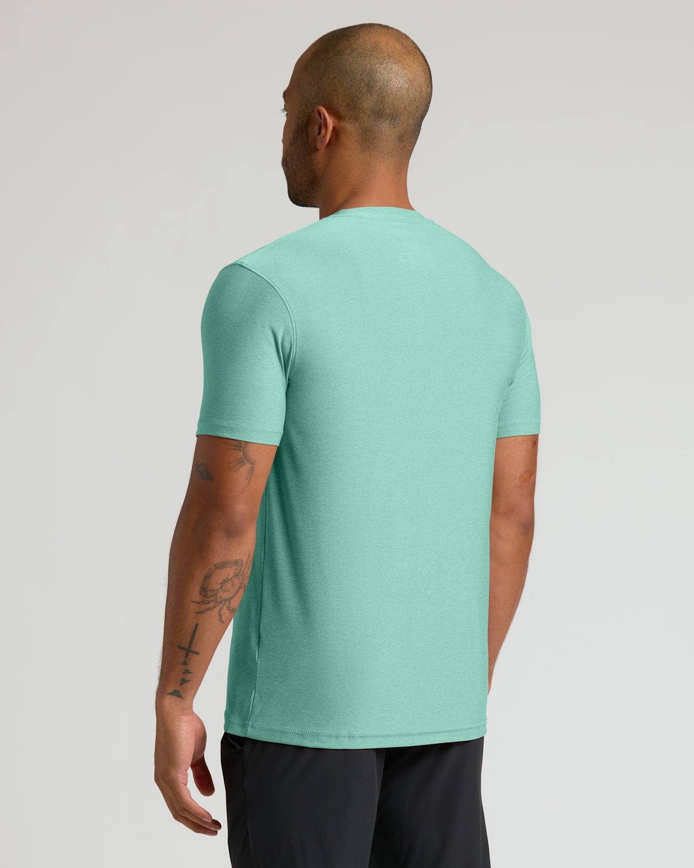 A man viewed from the back wearing a short-sleeve crewneck t-shirt in a heathered mint green color, paired with black athletic pants, against a light gray studio background.