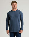 A man wearing a slate blue long-sleeve crewneck t-shirt and dark blue jeans, standing against a light gray studio background.