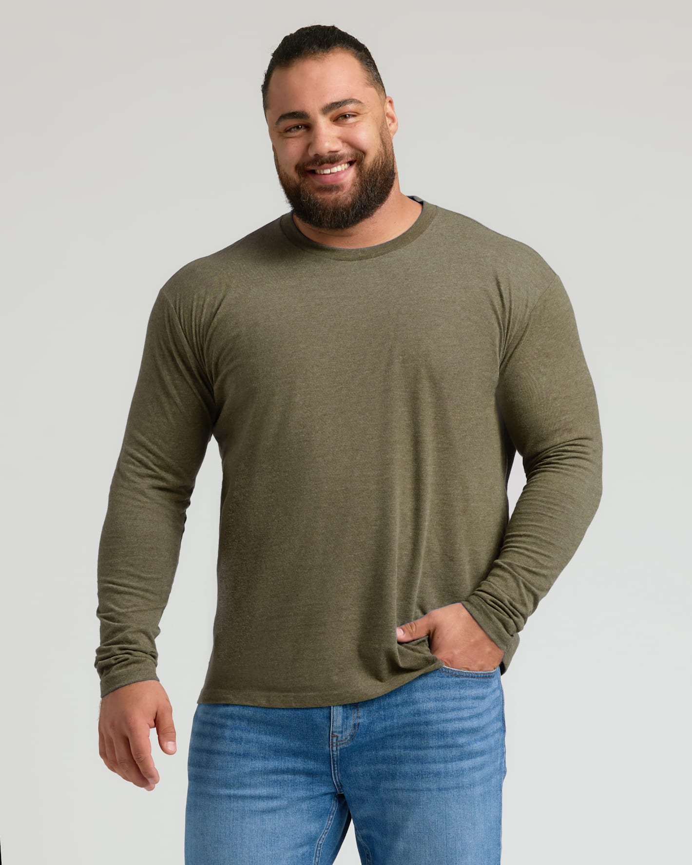 A smiling man with a beard wearing an olive green long-sleeve crewneck shirt and blue jeans, standing against a light gray studio background.