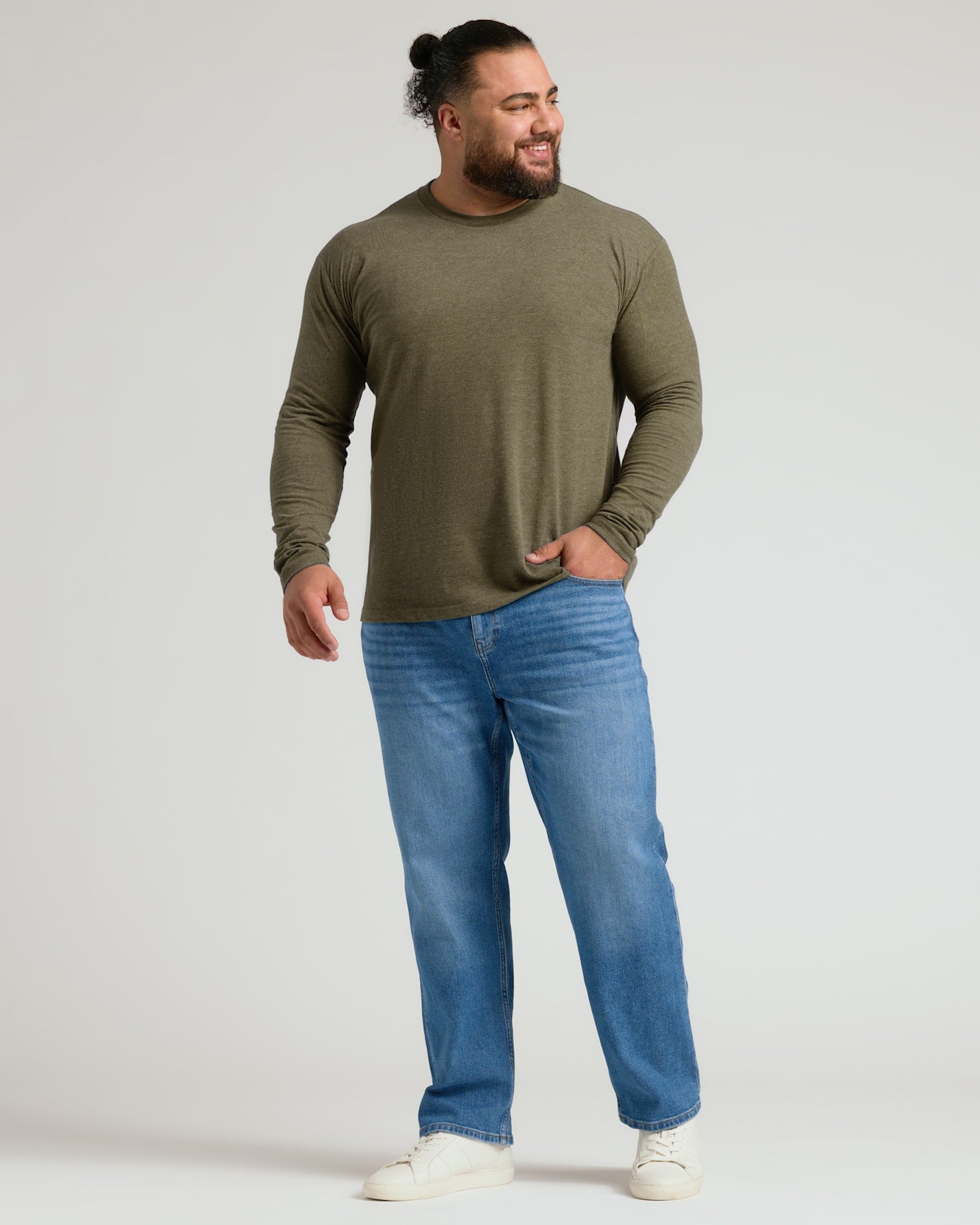 A man wearing an olive green long-sleeve crewneck shirt, blue jeans, and white sneakers, standing against a light gray studio background.