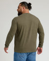 A man with a beard and a bun, viewed from the back, wearing an olive green long-sleeve crewneck shirt and blue jeans against a light gray studio background.