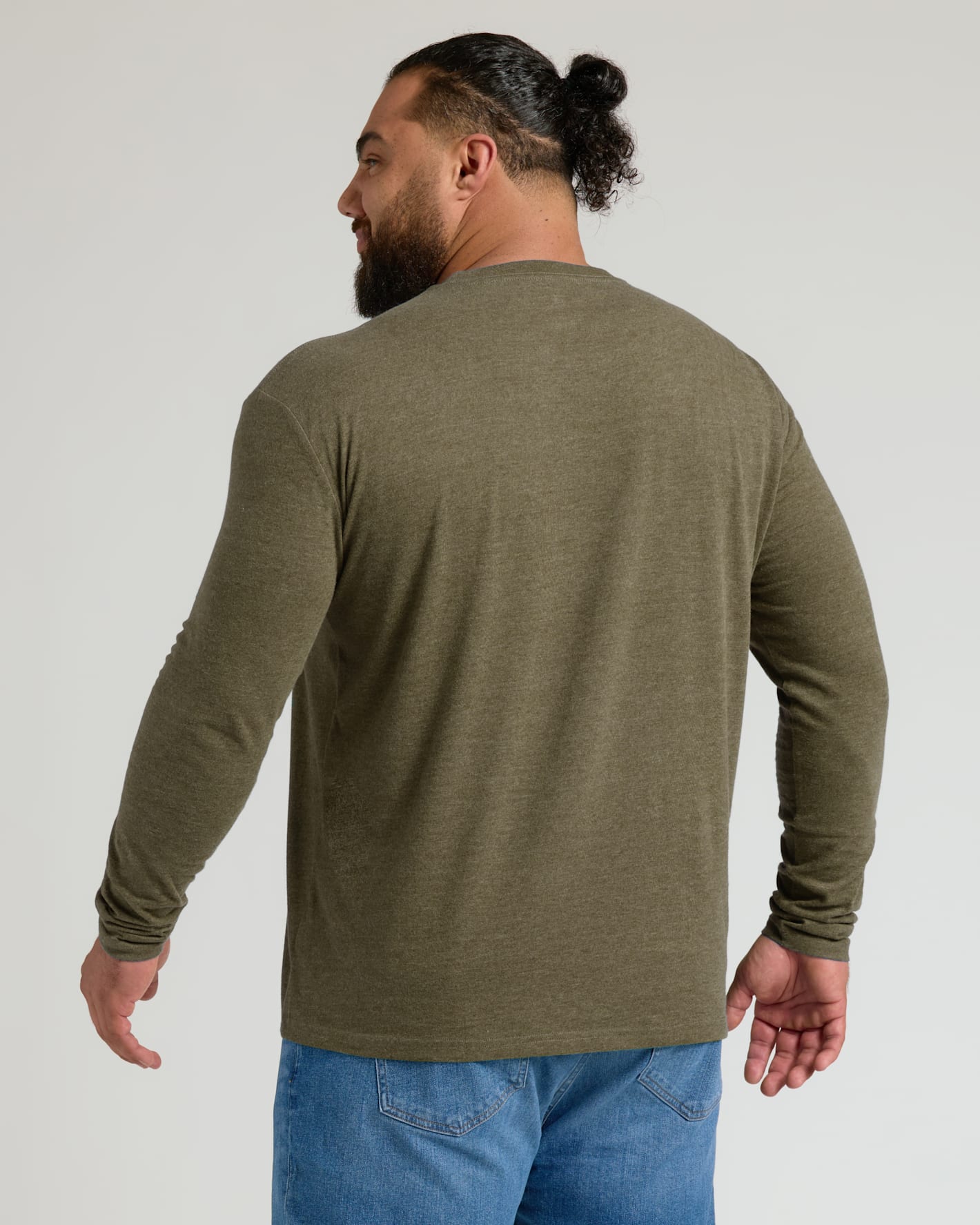 A man with a beard and a bun, viewed from the back, wearing an olive green long-sleeve crewneck shirt and blue jeans against a light gray studio background.
