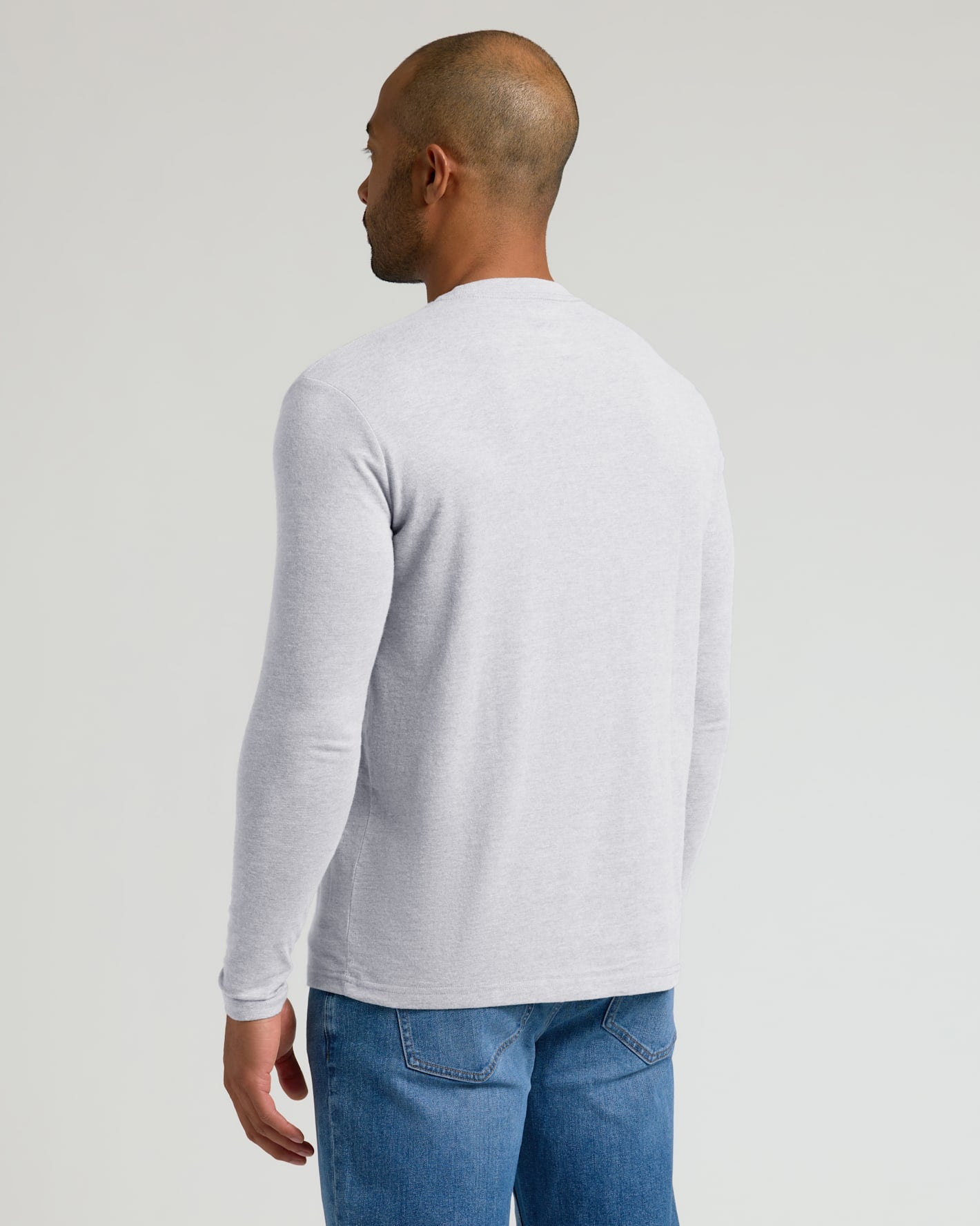 A man shown from the back wearing a light gray long-sleeve crewneck shirt and blue jeans, photographed against a solid light gray studio background.