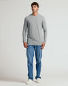 A man modeling a light gray long-sleeve crewneck t-shirt and blue jeans against a plain light gray studio background.