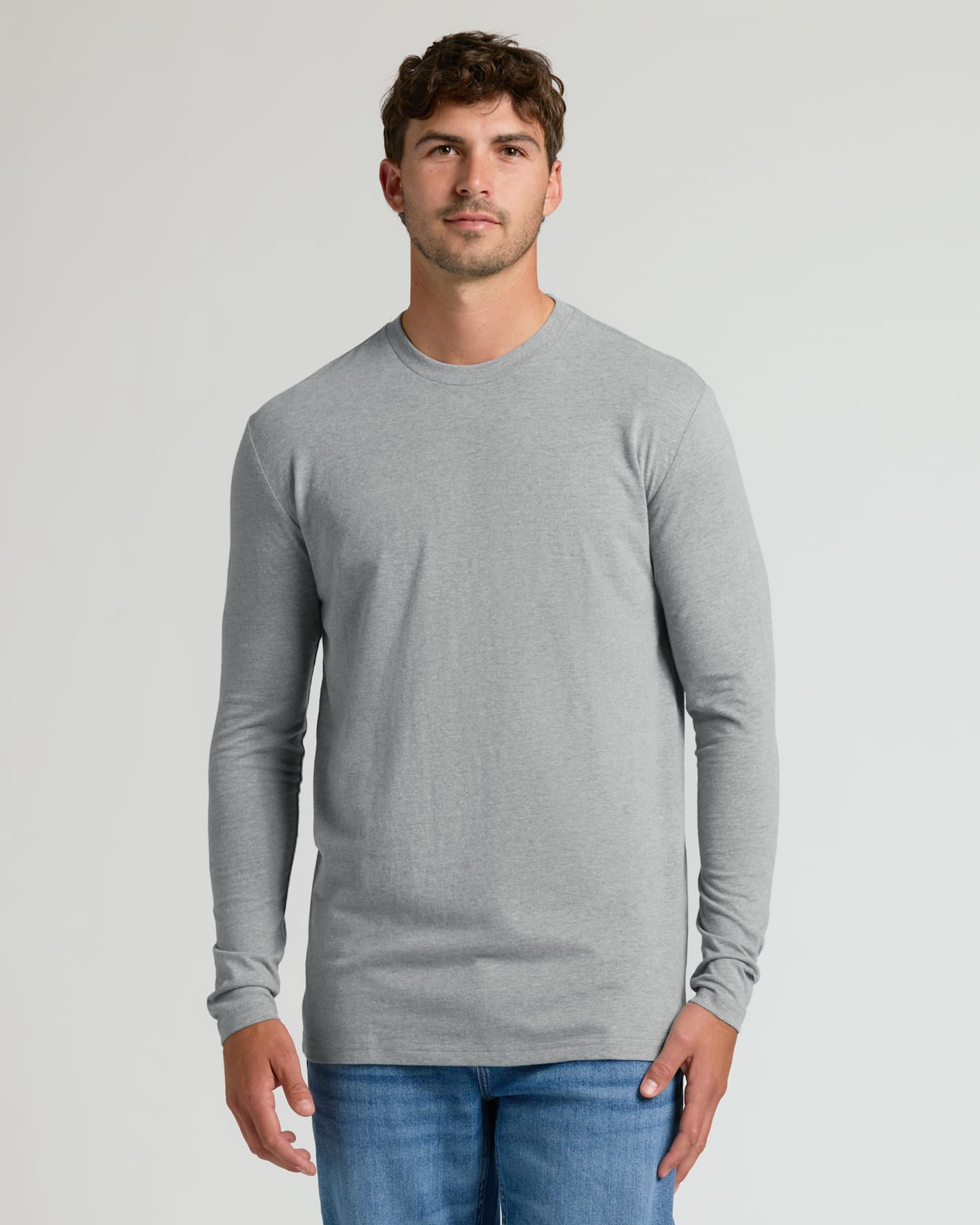 A man wearing a light gray long-sleeve crewneck t-shirt and blue jeans, standing against a light gray studio background.