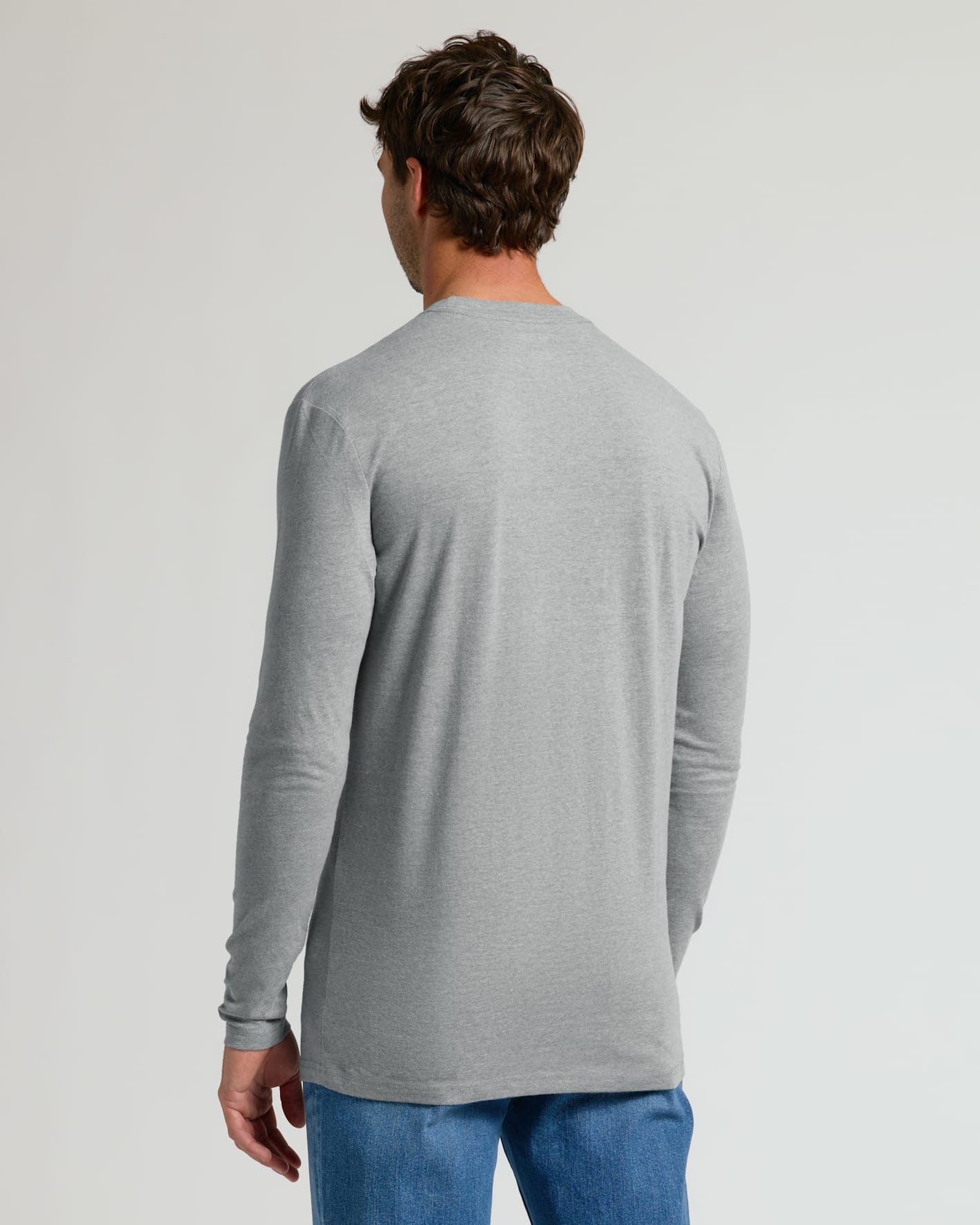 A man viewed from the back wearing a light gray long-sleeve t-shirt and blue jeans, standing against a plain light gray studio background.