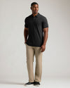 A man modeling a black short-sleeve polo shirt paired with beige trousers and black shoes, standing against a light gray studio background.
