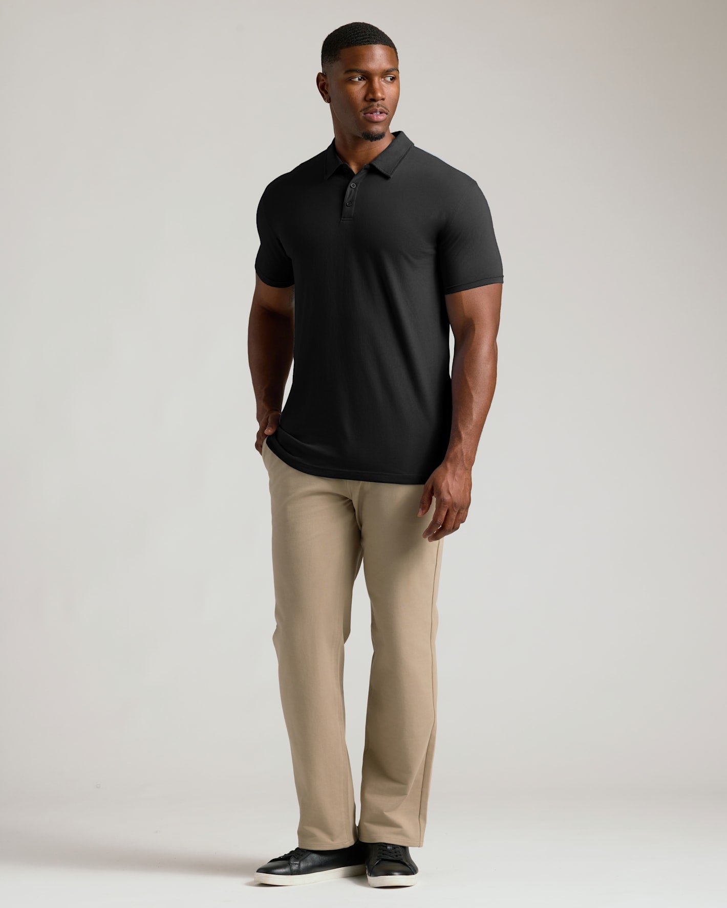A man modeling a black short-sleeve polo shirt paired with beige trousers and black shoes, standing against a light gray studio background.