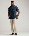 A man modeling a short-sleeve polo shirt in navy blue, paired with beige trousers and black slip-on shoes, standing against a light gray studio background.