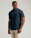 A man wearing a short-sleeve polo shirt in dark navy blue, paired with light beige pants, standing against a solid light gray studio background.