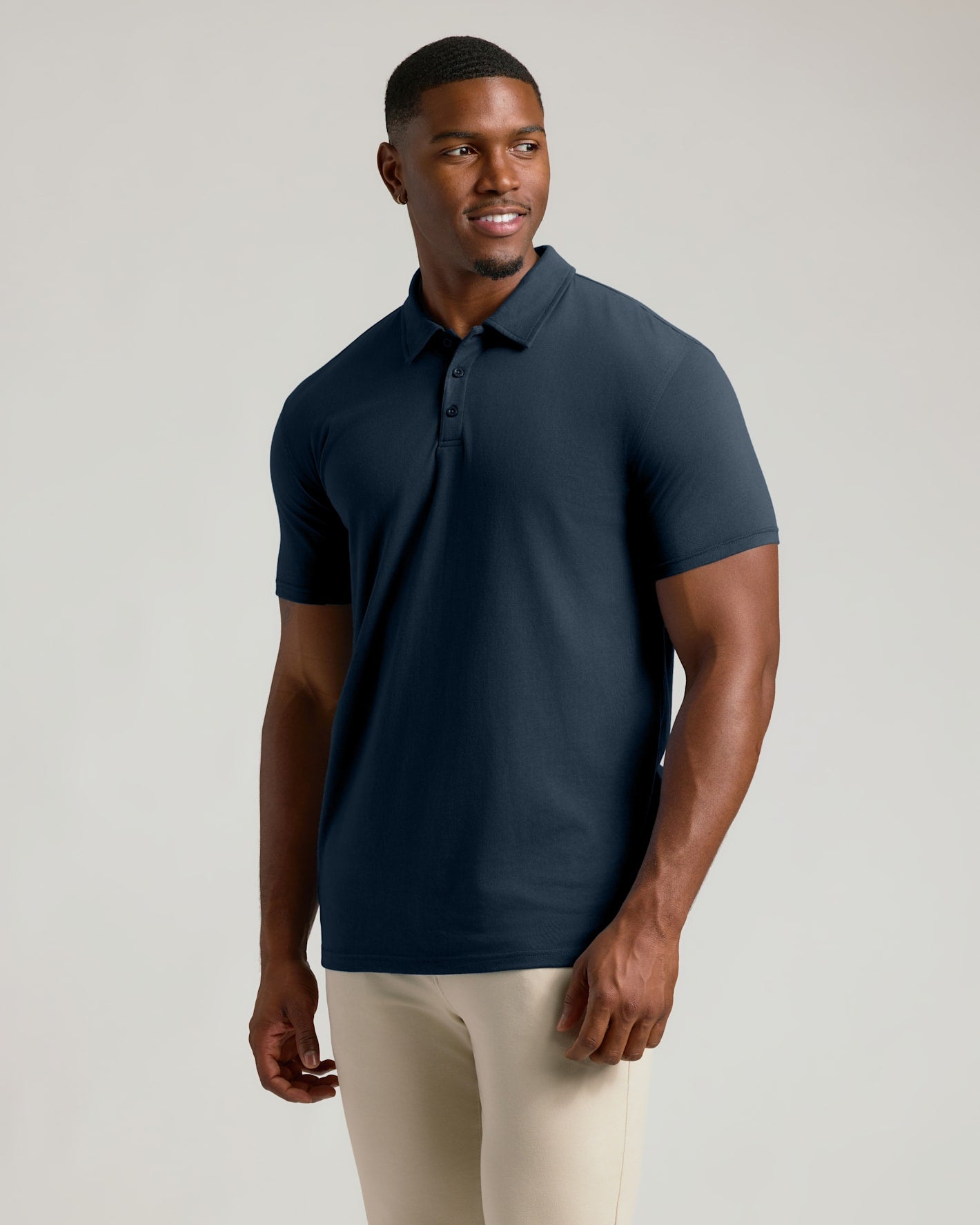 A man wearing a short-sleeve polo shirt in dark navy blue, paired with light beige pants, standing against a solid light gray studio background.
