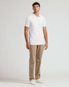 A man modeling a white short-sleeve polo shirt, paired with beige trousers and white sneakers, standing against a light gray studio background.