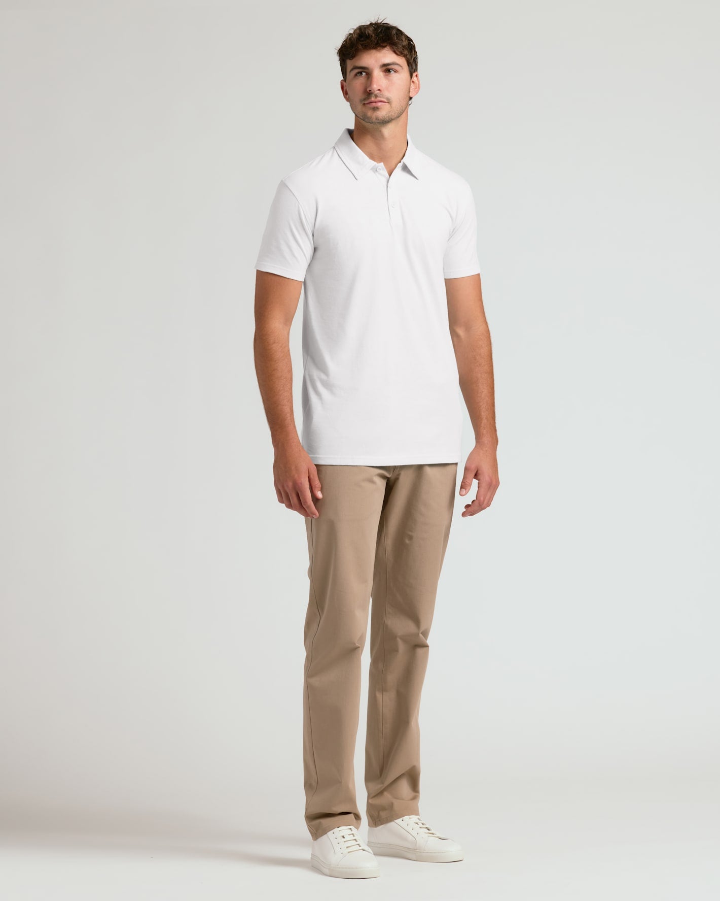A man modeling a white short-sleeve polo shirt, paired with beige trousers and white sneakers, standing against a light gray studio background.