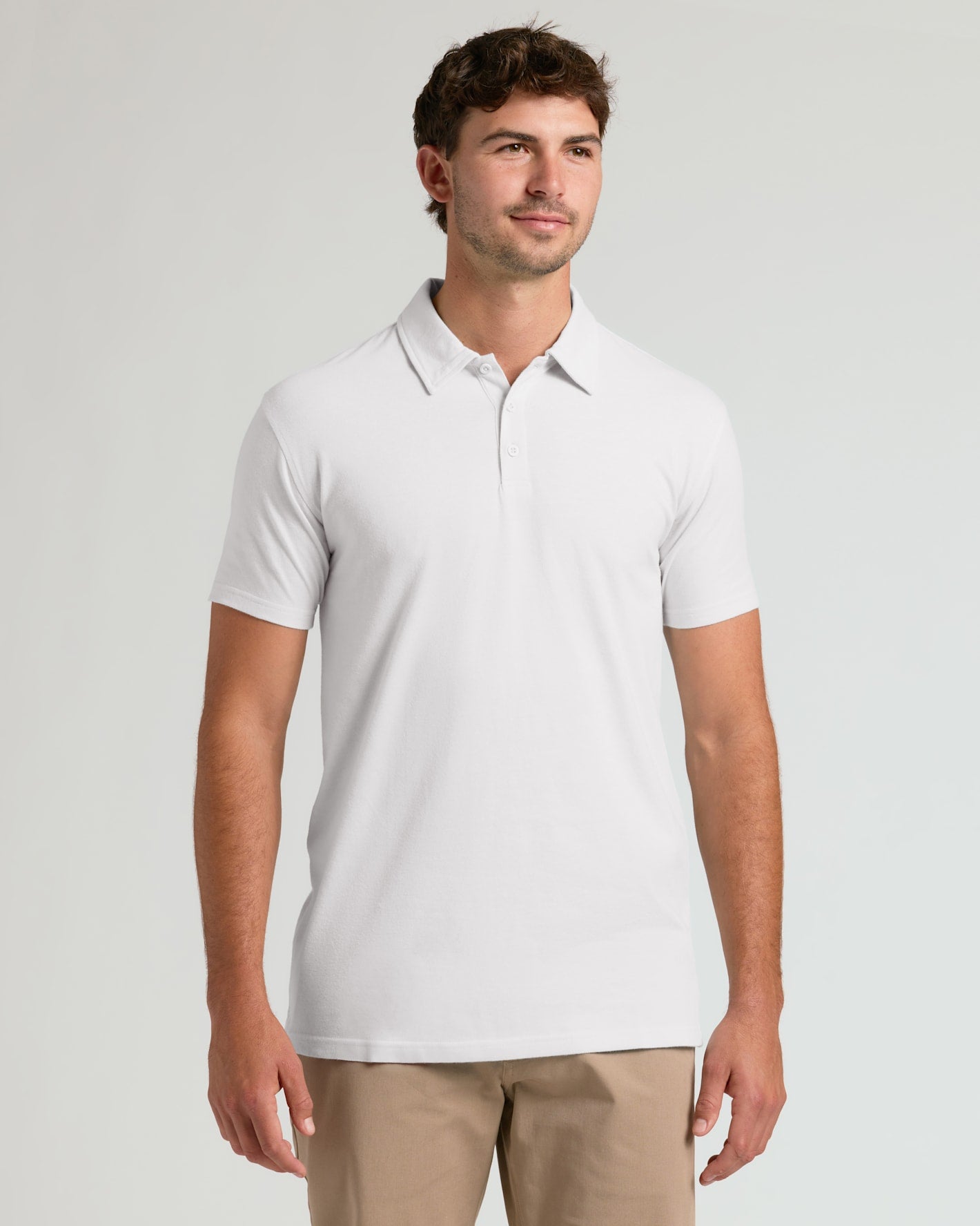 A man wearing a white short-sleeve polo shirt and khaki pants, standing against a light gray studio background.
