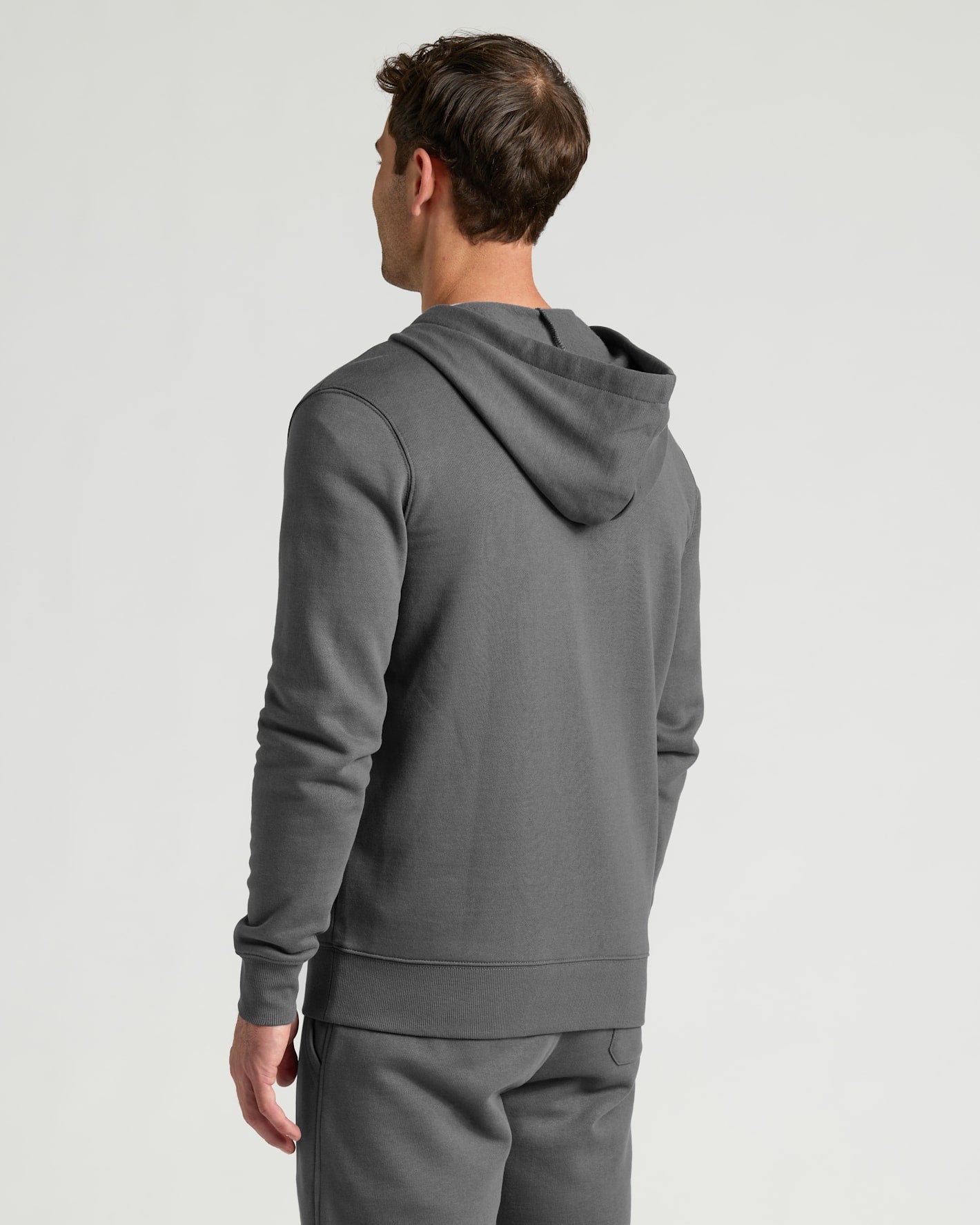 A man wearing a dark gray pullover hoodie and matching sweatpants, shown from a rear three-quarter view against a light gray studio background.