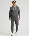 A man modeling a dark gray ribbed hoodie and matching jogger pants set, paired with white sneakers, against a light gray studio background.