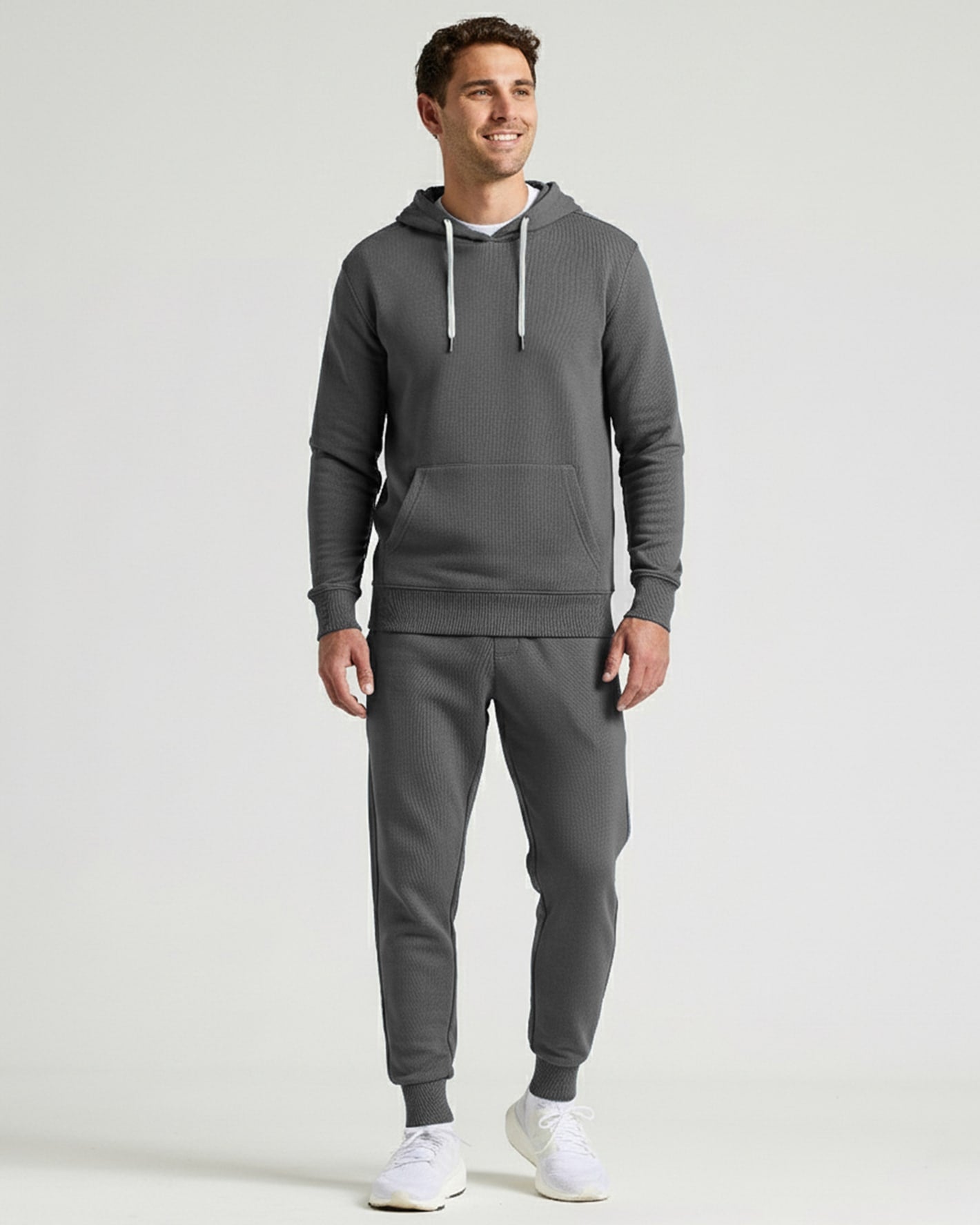 A man modeling a dark gray ribbed hoodie and matching jogger pants set, paired with white sneakers, against a light gray studio background.