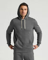 A man wearing a dark gray pullover hoodie with light beige drawstrings and matching gray sweatpants, standing against a light gray studio background.