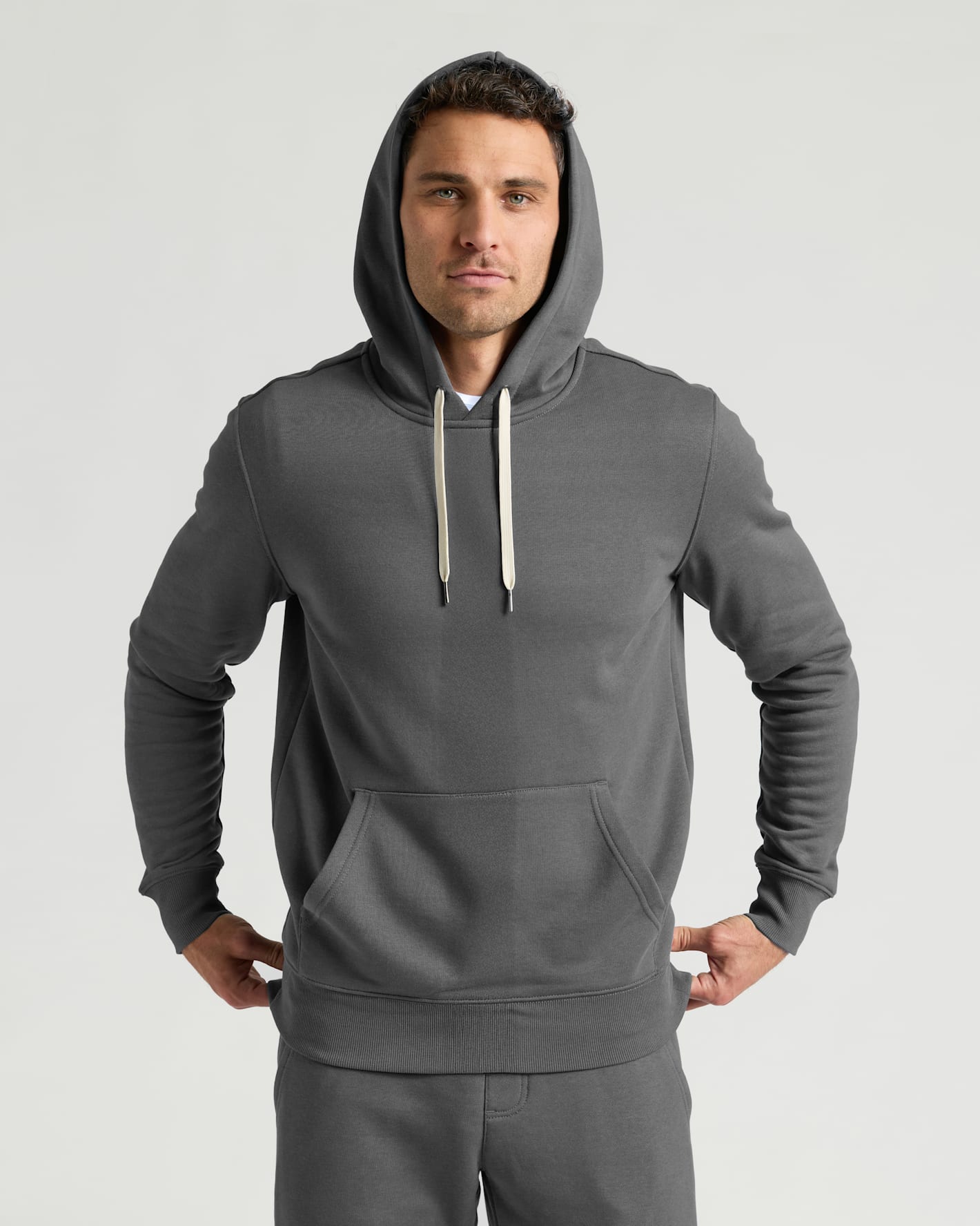 A man wearing a dark gray pullover hoodie with light beige drawstrings and matching gray sweatpants, standing against a light gray studio background.