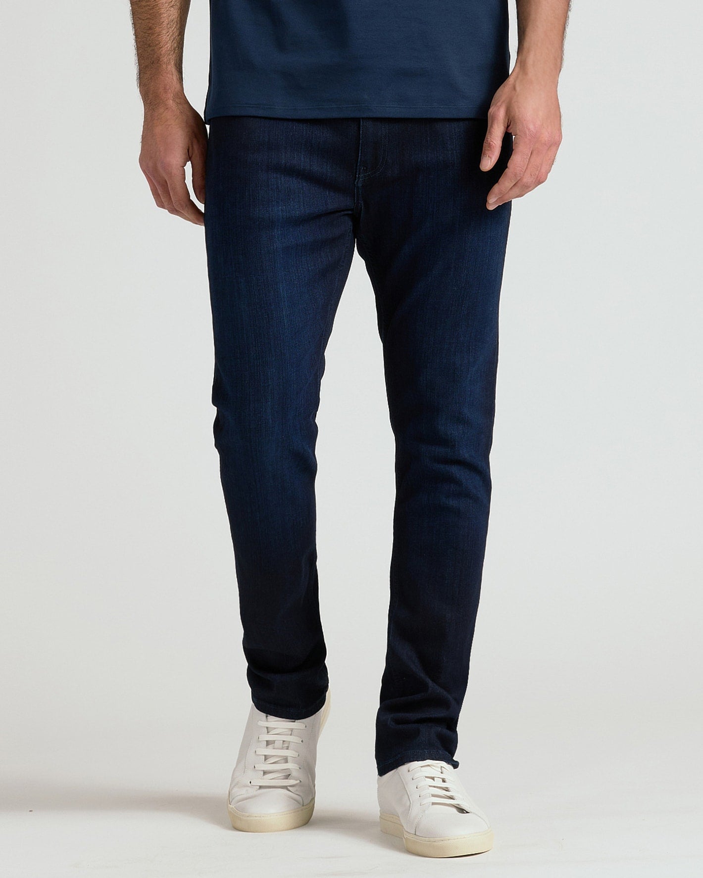 A person wearing slim-fit dark blue denim jeans and white sneakers, shown from the waist down against a plain light gray studio background.