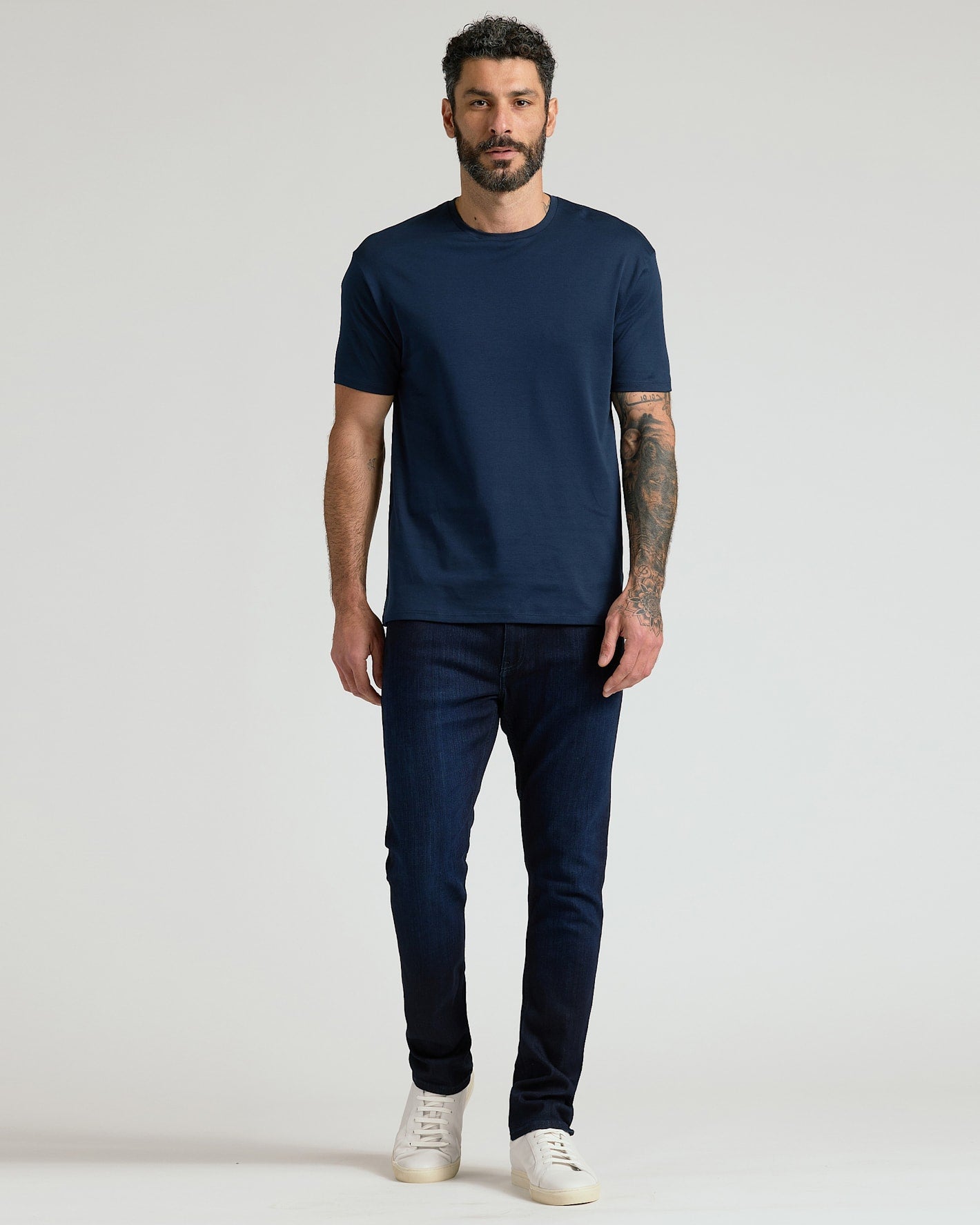 A man modeling a dark navy blue crew-neck t-shirt and dark navy blue jeans, paired with white sneakers, against a plain light gray studio background.