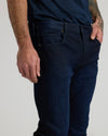Close-up of a person wearing dark indigo denim jeans with a navy t-shirt, showing the front pocket and waistband details against a light gray background.