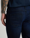Close-up back view of a person wearing dark indigo denim jeans, showing the back pocket and waistband, with a tattooed arm visible at the left edge against a light gray studio background.