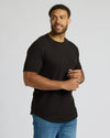 Man wearing a black True Classic Men's Tall Heavyweight Curved Hem Crew – Premium Heavyweight Cotton Blend, Extended Length, Athletic Fit, Structured Everyday T-Shirt with blue jeans against a plain background.