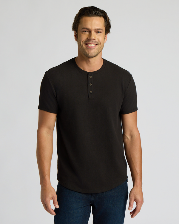 Man wearing a black Heavyweight Curved Hem Henley 3-Pack shirt with short sleeves and buttoned placket, paired with dark blue jeans, smiling against a plain background.