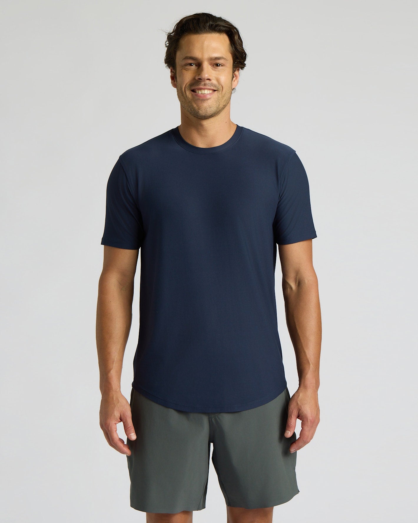 Man wearing a navy blue Active Curved Hem Crew Tee - Men's Moisture Wicking Performance T-Shirt paired with gray shorts, standing and smiling against a plain background.