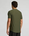 A man viewed from the back wearing a short-sleeve olive green athletic t-shirt and black pants, set against a plain light gray studio background.