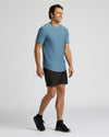 A man wearing a light blue short-sleeve athletic t-shirt, black athletic shorts, and black sneakers, standing against a light gray studio background.