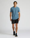 A man modeling a slate blue crewneck t-shirt and black athletic shorts against a light gray studio background.