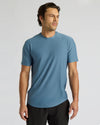 A man wearing a short-sleeve crew-neck athletic t-shirt in a muted slate blue color, paired with black athletic pants, photographed against a neutral light gray studio background.