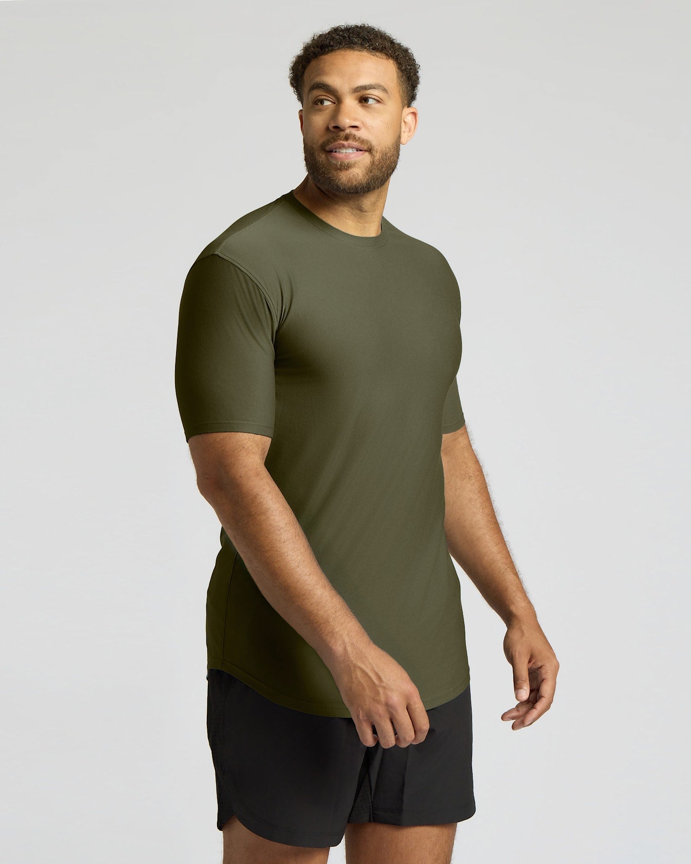 A man wearing a fitted short-sleeve athletic t-shirt in olive green, paired with black shorts, photographed against a light gray studio background.