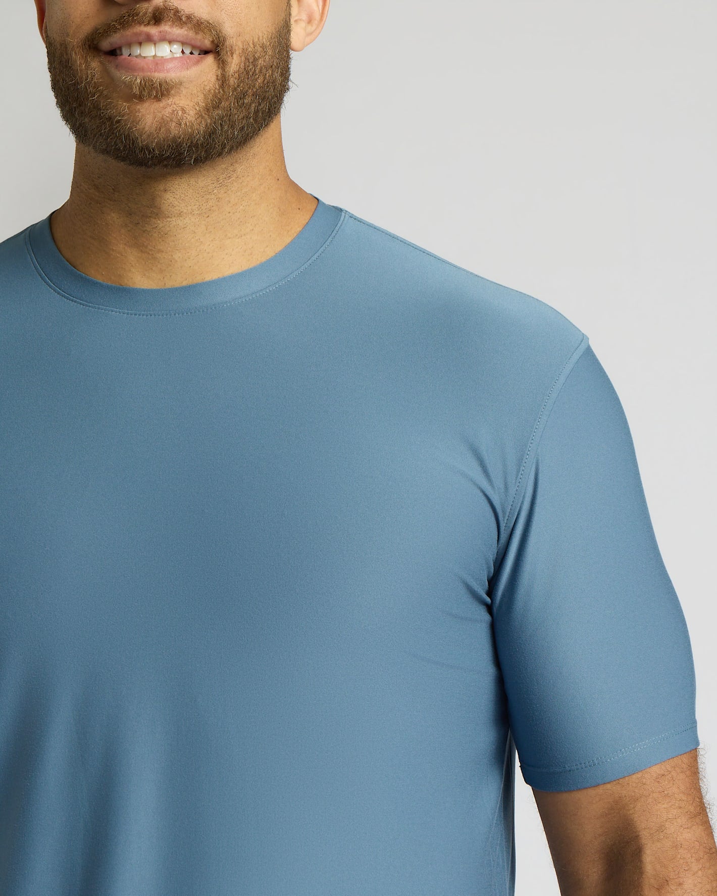 A close-up of a man wearing a short-sleeve crew-neck athletic t-shirt in a dusty slate blue color, shown from the chin down against a light gray background.