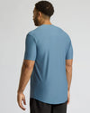 A man viewed from the back wearing a short-sleeve athletic t-shirt in a dusty blue color, paired with black pants, against a light gray studio background.