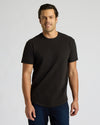 Man wearing a black True Classic Men's Heavyweight Curved Hem Crew – Premium Heavyweight Cotton Blend, Modern Athletic Fit, Structured Everyday T-Shirt standing and smiling against a plain background.