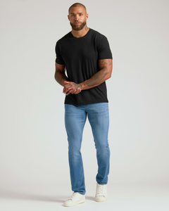 True ClassicAthletic man wearing light blue Slim Comfort Stretch Jeans 3-Pack paired with a black t-shirt and white sneakers against a plain background.